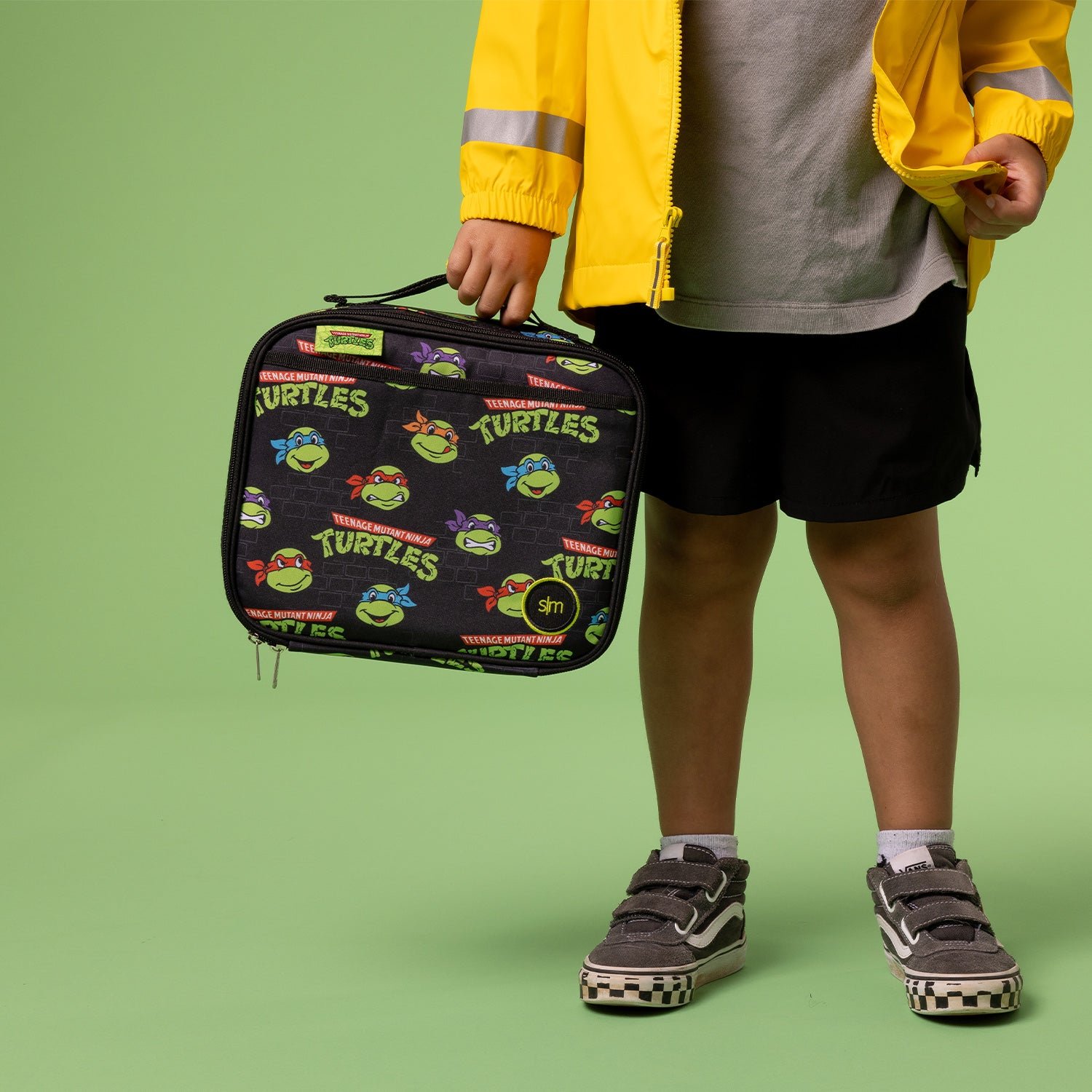 Child holding a Teenage Mutant Ninja Turtles lunchbox, wearing a yellow jacket, shorts, and sneakers.