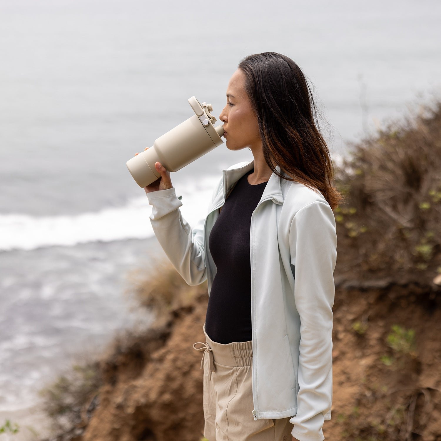 A woman drinking from a beige bottle near a seaside cliff.