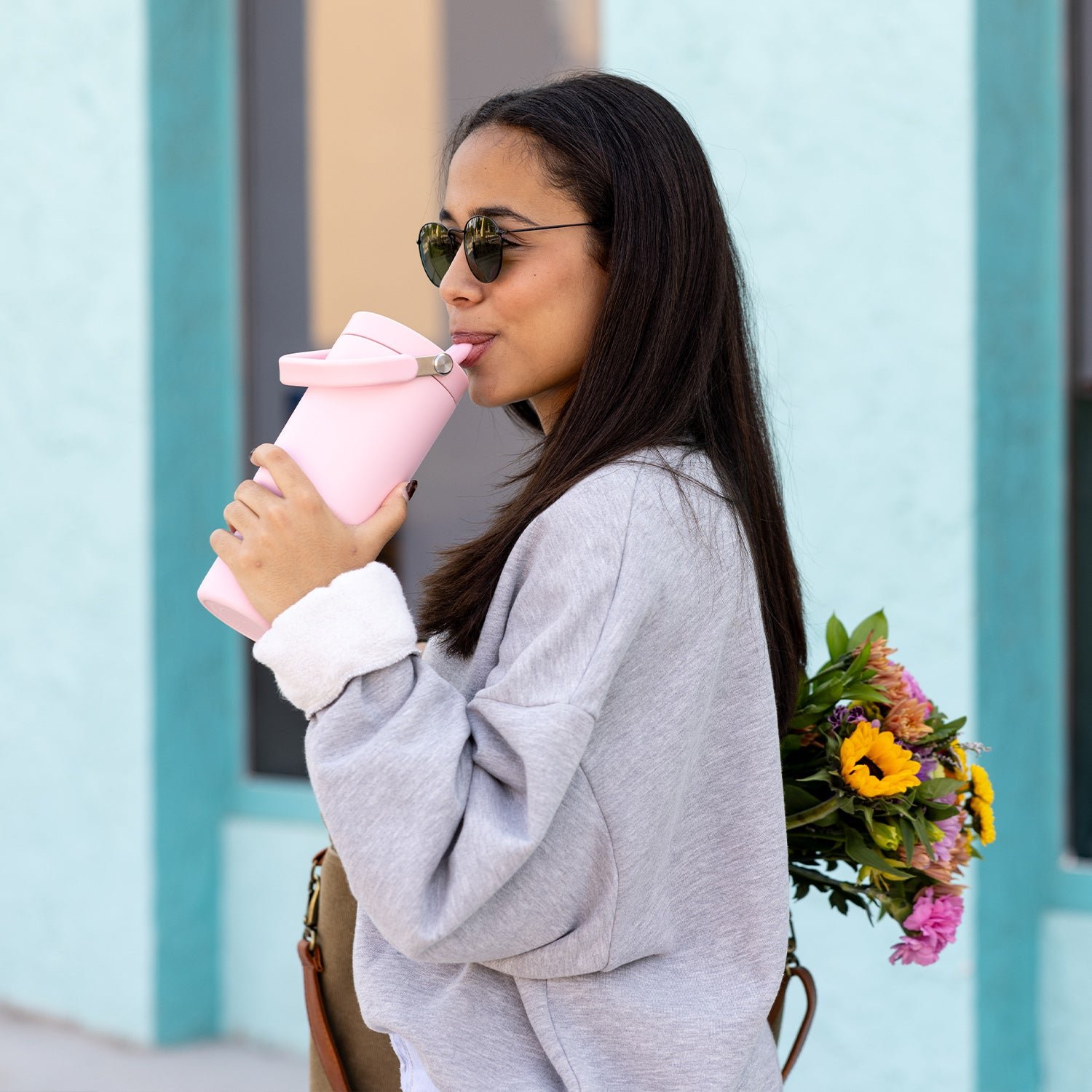 Woman drinking from a pink bottle, holding flowers, and wearing sunglasses and a gray sweatshirt.