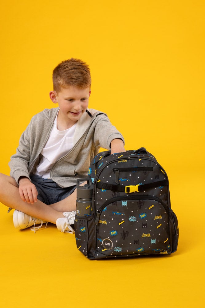 A child seated, reaching into a Batman-themed backpack, against a yellow background.